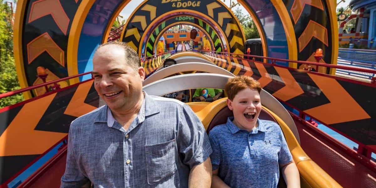 A man and a boy excitedly ride a vibrant roller coaster. The track features bold, colorful patterns creating a dynamic visual backdrop. Both are smiling widely, capturing the thrill and joy of the moment.