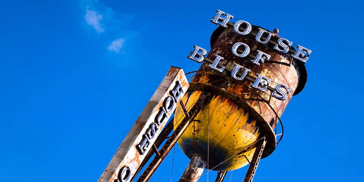 Tilted view of a rusty water tower with "House of Blues" in bold letters against a clear blue sky, reminiscent of Disney World lounges. Another smaller sign reads "House" vertically. The tower's weathered look adds vintage charm.