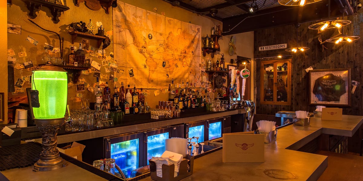 A cozy bar reminiscent of Disney World lounges, with illuminated shelves filled with various liquor bottles. A vintage map hangs on the wall, and a glowing green light decorates the counter. Above, warm pendant lights create a welcoming ambiance.