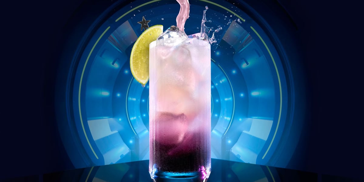 A vibrant cocktail splashes in a tall glass against a futuristic blue background, reminiscent of the magical ambiance of Disney World lounges. The drink features a gradient from purple to pink and is garnished with a lime wedge on the rim. A star decoration brightly adorns the glass.