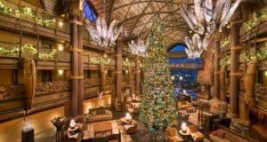 A grand hotel lobby decorated for the holidays with a towering Christmas tree adorned with lights and ornaments. Festive garlands wrap around the balconies and large, unique chandeliers hang from the ceiling. Comfortable seating areas surround the tree.