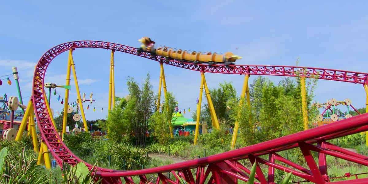 A colorful roller coaster with red tracks and yellow supports winds through a theme park. A train with excited passengers zooms along the curve. The surrounding area is lush with greenery and playful decorations. The sky is clear and blue.
