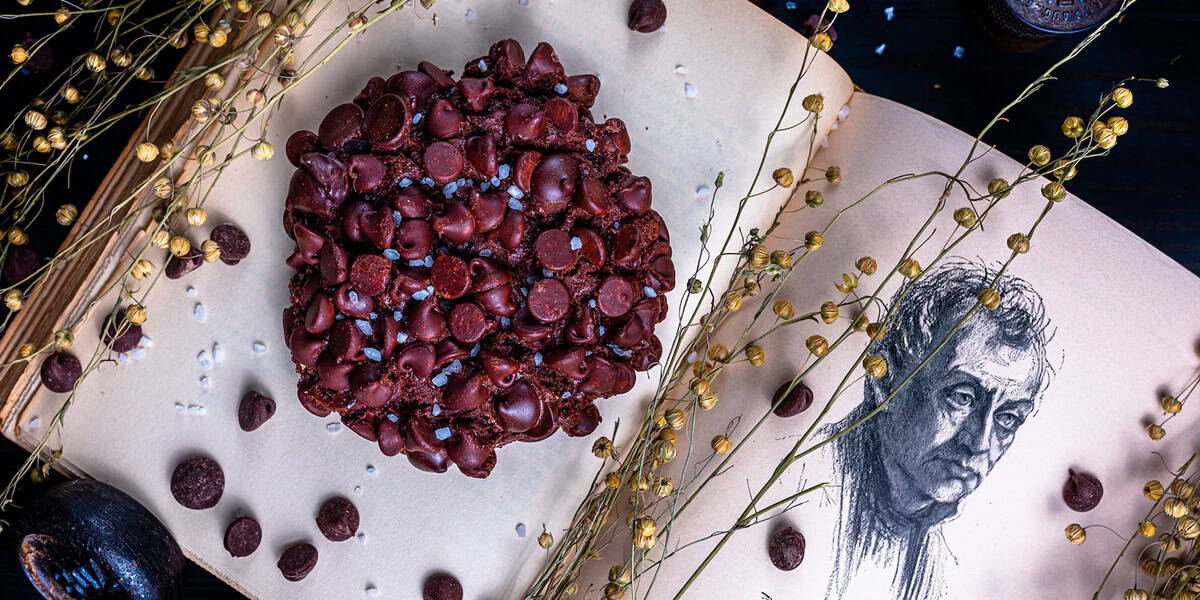 A large chocolate chip cookie rests on an open book next to a black-and-white drawing of a man’s face, surrounded by dried flowers and scattered chocolate chips—a scene reminiscent of cozy Disney Springs activities.