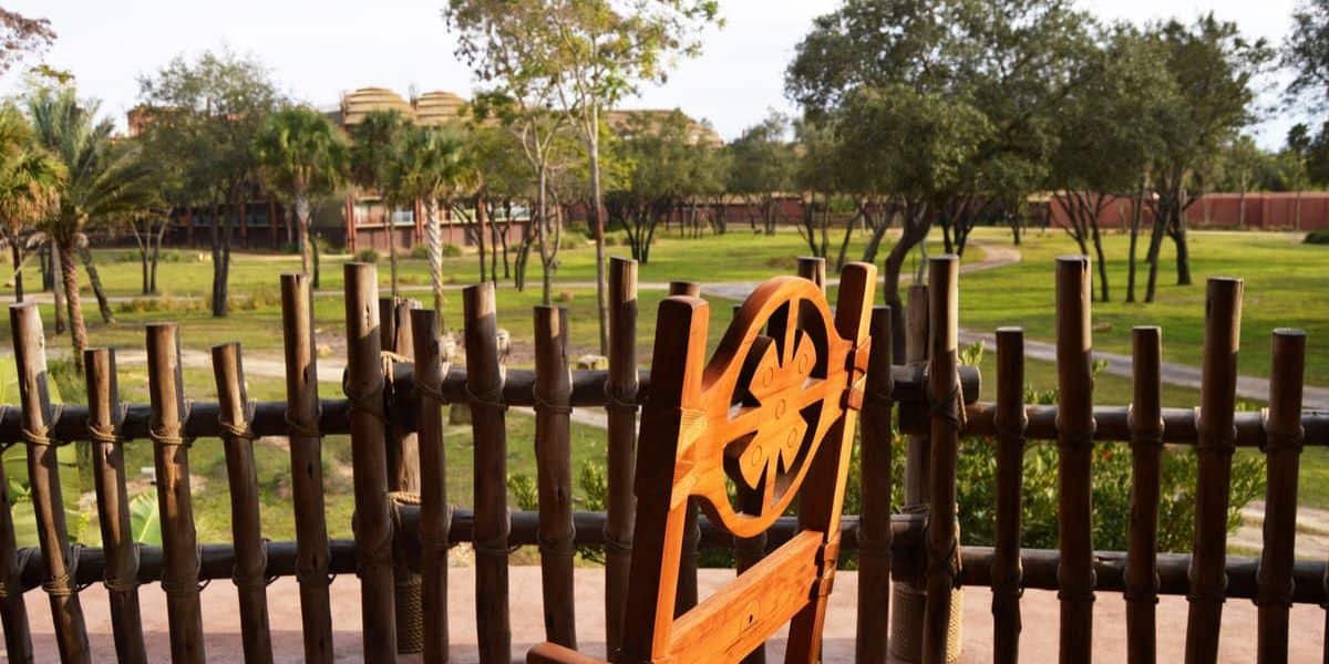 A wooden chair with a carved backrest overlooks a wide grassy field, enclosed by a rustic bamboo fence. Trees are scattered across the field, and buildings resembling Magical Disney Moments are visible in the distance under a clear sky.
