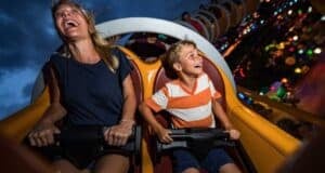 A woman and a young boy are seated on a roller coaster, both smiling and laughing with excitement. The ride is illuminated with colorful lights in the background, contrasting against a dark, cloudy sky.