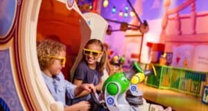 Two children wearing 3D glasses sit in a colorful amusement park ride, smiling and holding toy blasters. The background features vibrant lights and playful decor, creating a fun and lively atmosphere.