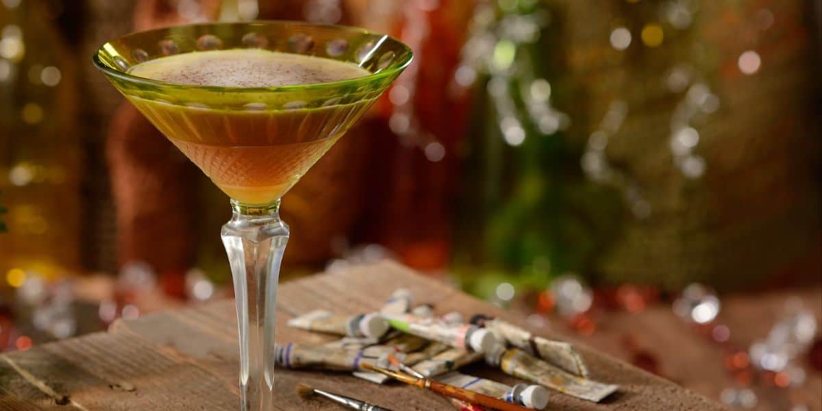 A martini glass filled with a layered cocktail sits on a rustic wooden surface. Several small paint tubes and brushes are scattered nearby. The background features colorful, blurred lights, creating a warm, artistic ambiance.