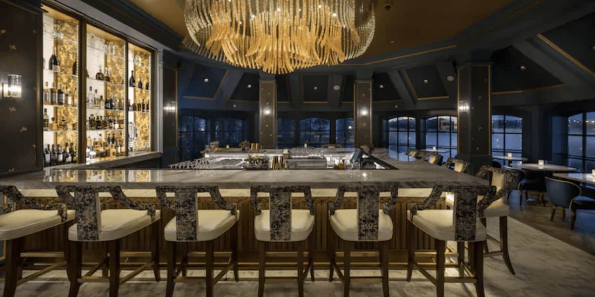 A luxurious bar with sleek, modern decor, featuring a long marble counter, elegant bar stools, and a large, ornate chandelier. The background showcases shelves of bottles illuminated with warm lighting, set against a dark, sophisticated interior.