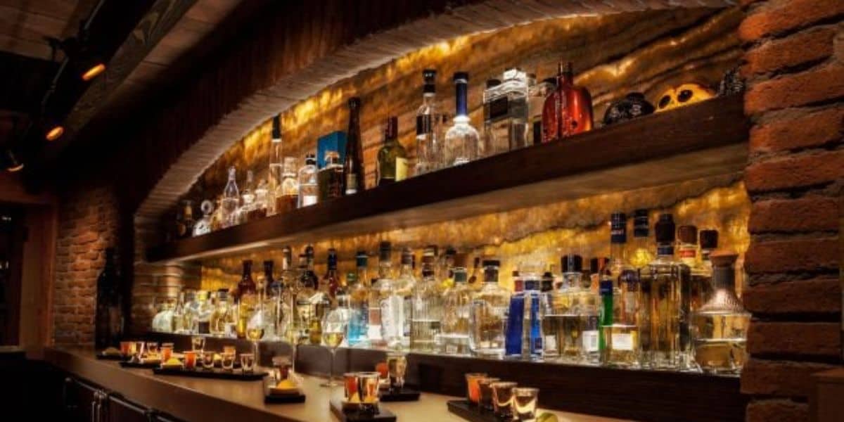 A dimly lit bar with a brick arch and shelves filled with various liquor bottles. Shot glasses are neatly arranged on the counter beneath warm, ambient lighting.