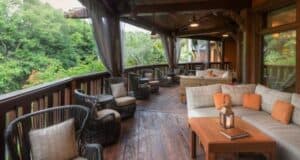 A cozy outdoor patio with wooden flooring, featuring cushioned sectional sofas and wicker chairs with pillows. A wooden coffee table with a lantern sits in the foreground. The patio overlooks lush greenery and is partially covered by a wooden roof.