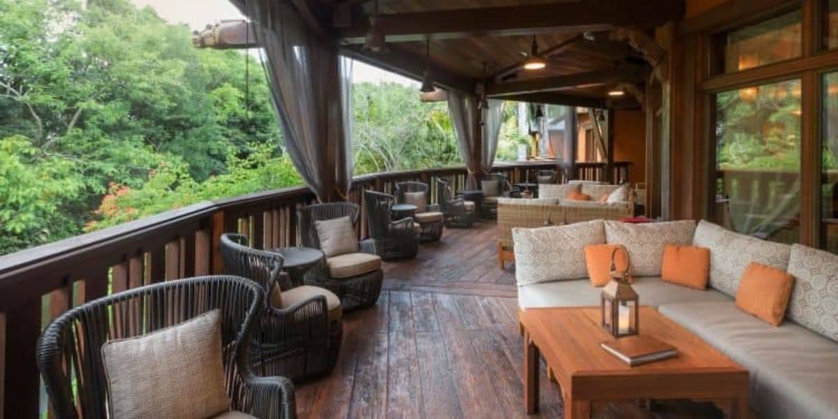 A cozy outdoor patio with wooden flooring, featuring cushioned sectional sofas and wicker chairs with pillows. A wooden coffee table with a lantern sits in the foreground. The patio overlooks lush greenery and is partially covered by a wooden roof.