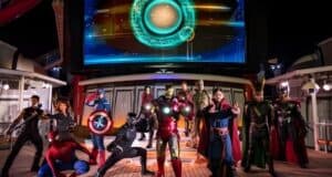 A group of actors dressed as various superheroes, including ones in red, blue, and green costumes, pose dramatically under a large screen displaying a glowing circular symbol. The scene is set in a futuristic, well-lit environment.