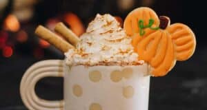A cozy white mug with polka dots is filled with a swirl of whipped cream, topped with a dusting of cinnamon, two rolled wafer cookies, and a pumpkin-shaped cookie. The background is softly blurred with warm lights.