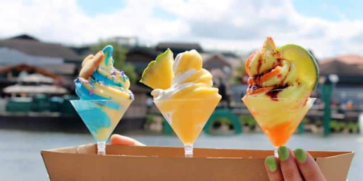 Three colorful ice cream cones with unique toppings are held up against a waterfront backdrop. The ice creams feature blue, yellow, and orange swirls topped with cookies, fruit slices, and drizzle, respectively. The background shows a calm water body and buildings.