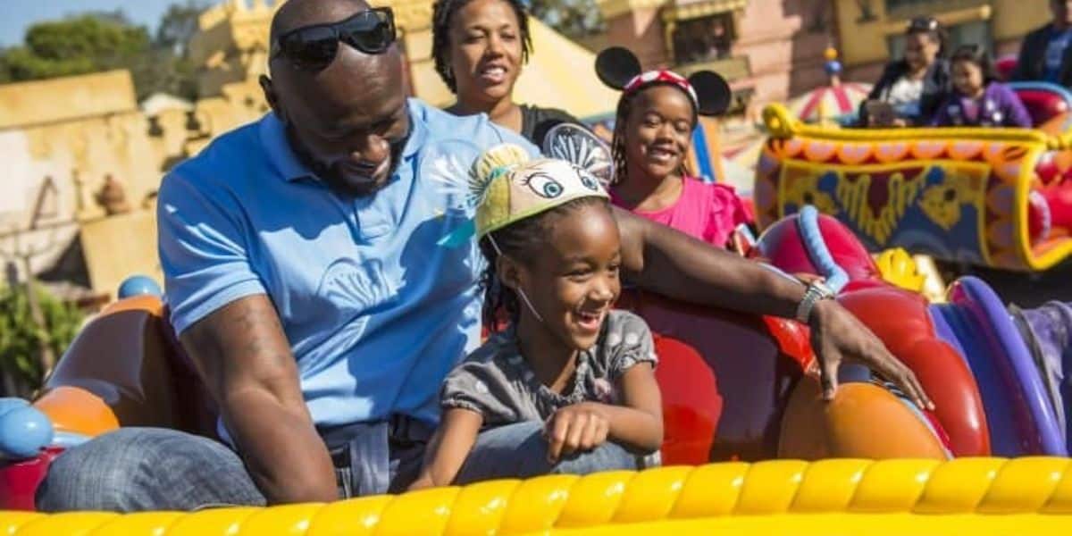 A family enjoys a ride on a colorful amusement park attraction. A father in sunglasses and a blue shirt sits beside his smiling daughter wearing a Disney-themed hat. Other park visitors are visible in the background.