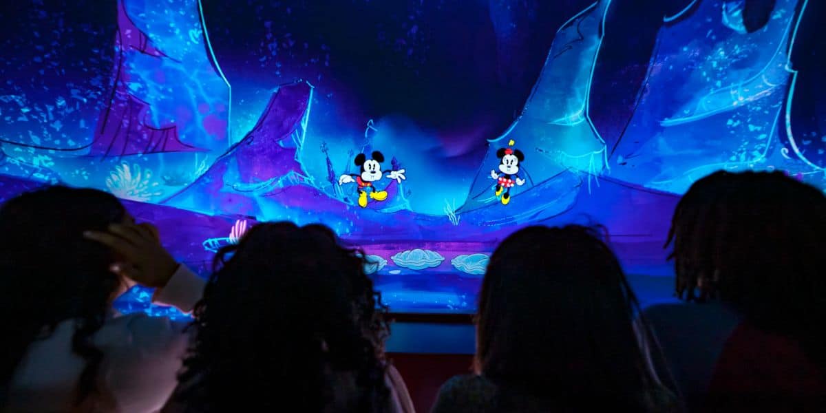 A group of people are watching an animated scene featuring two iconic cartoon characters on a colorful stage. The backdrop displays vibrant blue and purple hues with abstract shapes.
