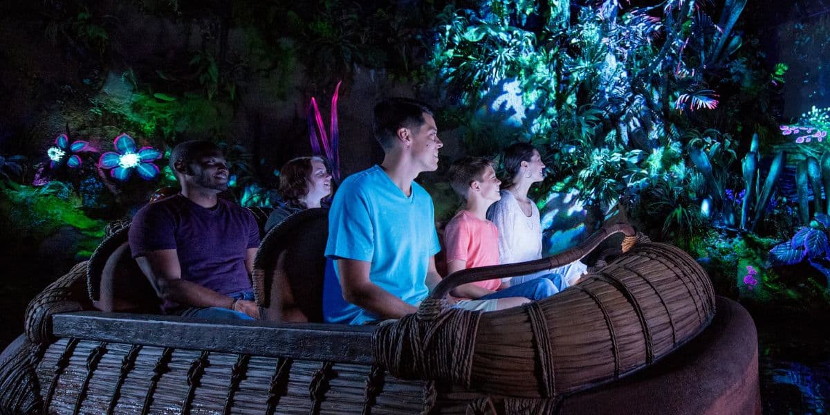 A group of people sit in a boat on an indoor ride, surrounded by colorful, glowing plants and futuristic, luminescent foliage. They look ahead, appearing engaged and entertained by the immersive environment.
