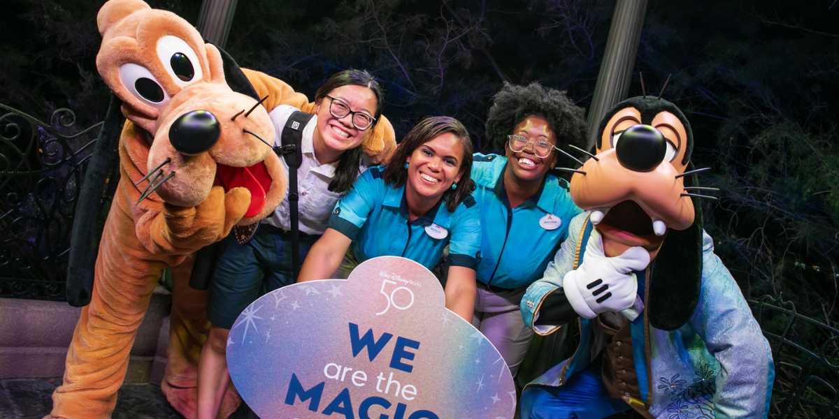 A group photo of three Disney cast members standing with Pluto and Goofy. The smiling cast members are holding a circular sign that reads, "We are the magic." They are outdoors, with trees and lights in the crisp background, truly showing what it means to be part of the Disney enchantment.