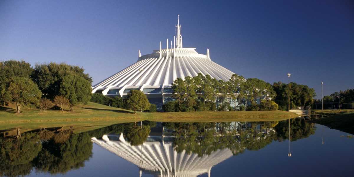 The image shows a futuristic, white, conical building with a pointed spire on top, reminiscent of Space Mountain in the Magic Kingdom. Surrounded by greenery and trees, the structure is reflected in a calm body of water in the foreground under a clear blue sky.