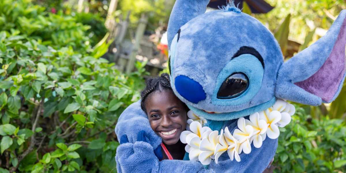 A person is smiling and posing outdoors with a large, plush costumed character resembling Stitch from the animated movie "Lilo & Stitch." The Disney cast member is wearing a white flower lei, and lush greenery is visible in the background.