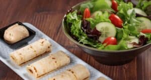 A fresh salad with mixed greens, cucumber slices, and cherry tomatoes sits in a bowl, perfect for healthy eating. Next to it, three rolled flatbreads lie on a rectangular plate with a small dish of dipping sauce. It's like dining at Walt Disney World right in your own home!