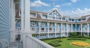 A view from a white balcony overlooking a courtyard with green grass and trimmed bushes, surrounded by a large, light blue, three-story building with Disney Resort Decor and many windows and balconies under a partly cloudy sky.