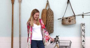 A woman with long blonde hair, wearing a white shirt and floral kimono, smiles while standing next to a vintage bicycle against a light blue wall adorned with nautical items inspired by Disney Resort Decor.