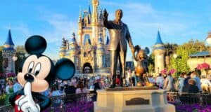 A statue of a man holding hands with a mouse character stands prominently in front of a fairy-tale castle. Amidst vibrant flowers and lush greenery, visitors enjoy the sunny day while considering Disney money-saving tips like arriving early for the best parking spots.