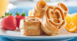A plate with waffles shaped like a cartoon character, accompanied by fresh strawberries and orange slices, sits atop a blue tablecloth—a delightful Disney Dining experience perfect for breakfast.