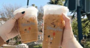 Two hands holding iced coffee cups with Disney Parks logos and playful Mickey Mouse silhouettes reflect the excitement of a sunny day. Could these refreshing delights be part of the elusive Disney Starbucks secret menu? Trees and a blurred green sign complete this magical moment.