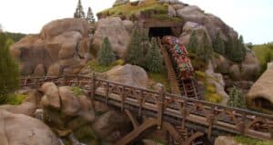 As the Seven Dwarfs Mine Train exits the tunnel and descends a hill at the theme park, it's surrounded by rock formations, small trees, and a wooden bridge. Thrilled riders eagerly raise their hands, fully immersed in the enchanting adventure.