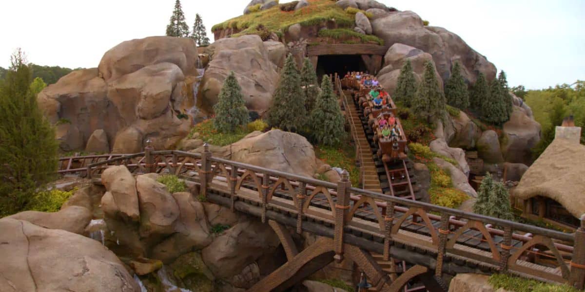As the Seven Dwarfs Mine Train exits the tunnel and descends a hill at the theme park, it's surrounded by rock formations, small trees, and a wooden bridge. Thrilled riders eagerly raise their hands, fully immersed in the enchanting adventure.