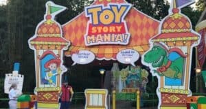 The entrance to Toy Story Midway Mania features colorful decorations with characters Jessie and Rex. Two staff members stand near the entrance under a large sign with flags and playful designs, inviting guests to the FastPass+ and Stand-by lines, ready to reveal the secret to achieving a high score.