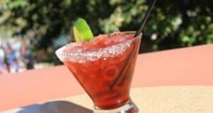 A red cocktail garnished with a lime wedge sits in a salt-rimmed glass on a sunny outdoor table, hinting at affordable drink prices. A black straw rests in the drink, with a blurred natural backdrop of greenery in the distance.
