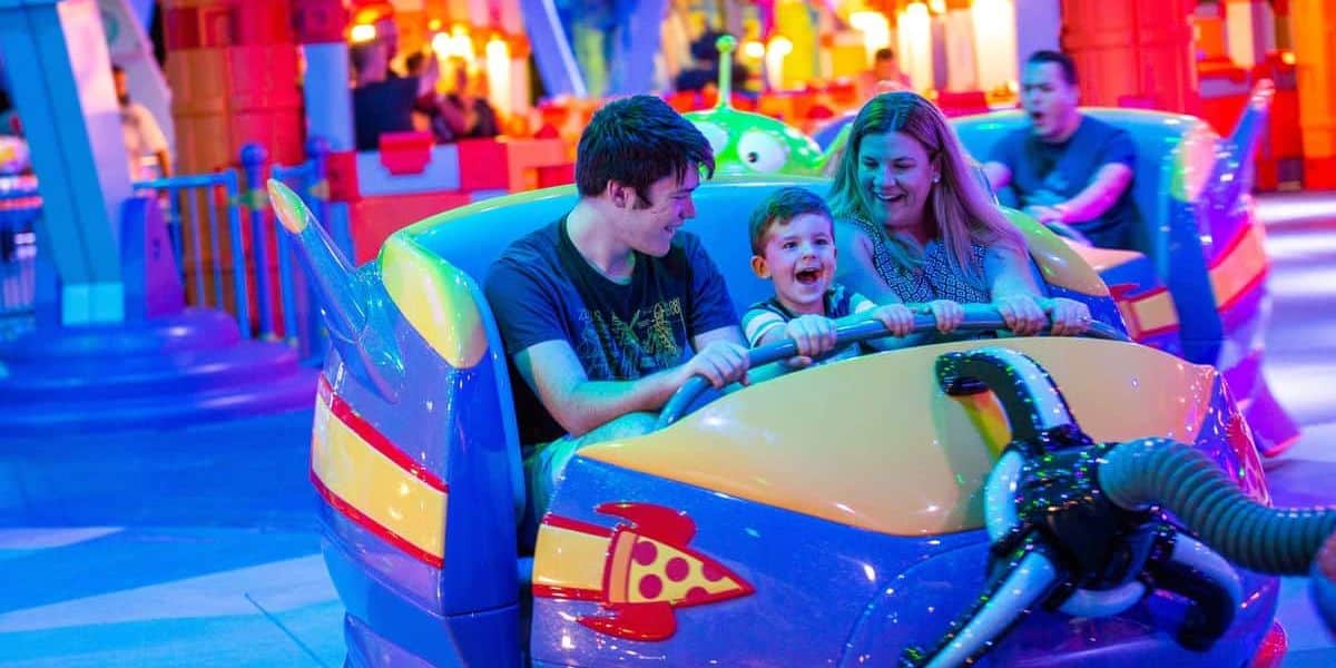A young family of three enjoys a colorful amusement park ride shaped like a spaceship at Walt Disney World. The child, sitting in the middle, laughs joyfully as the parents, seated on either side, smile and hold onto the ride. Bright, festive lights illuminate the background of this magical Disney attraction.