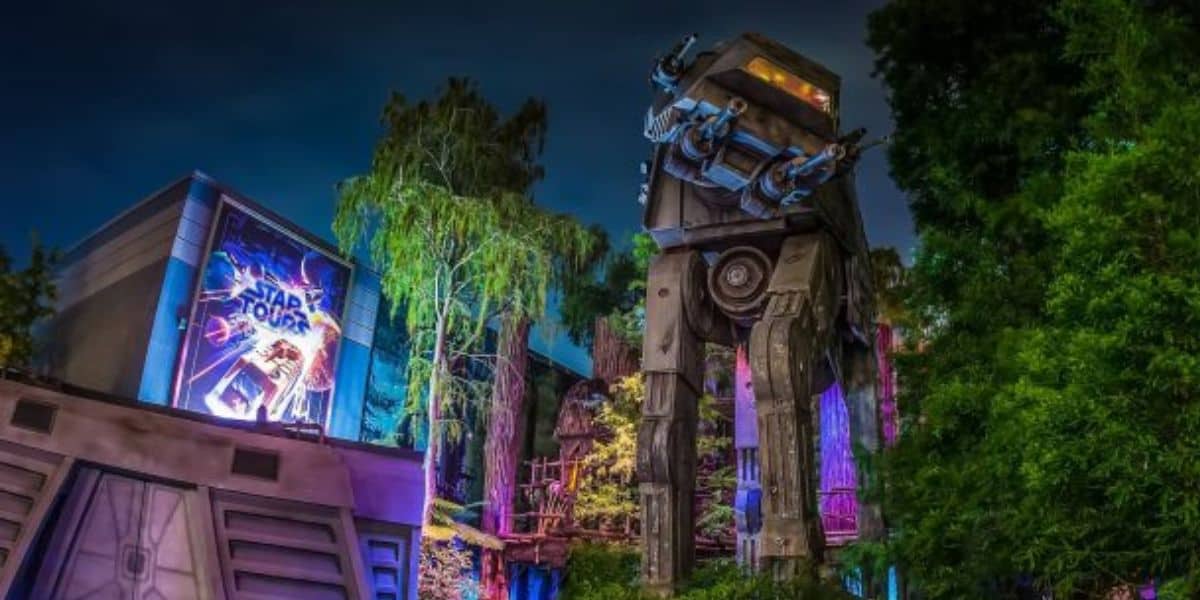 A nighttime view of a Star Wars-themed attraction at Walt Disney World features a large, illuminated AT-AT walker among trees. A brightly lit "Star Tours" sign is visible on a building to the left. The scene is enhanced with colorful lighting effects, showcasing the magic of Disney attractions.