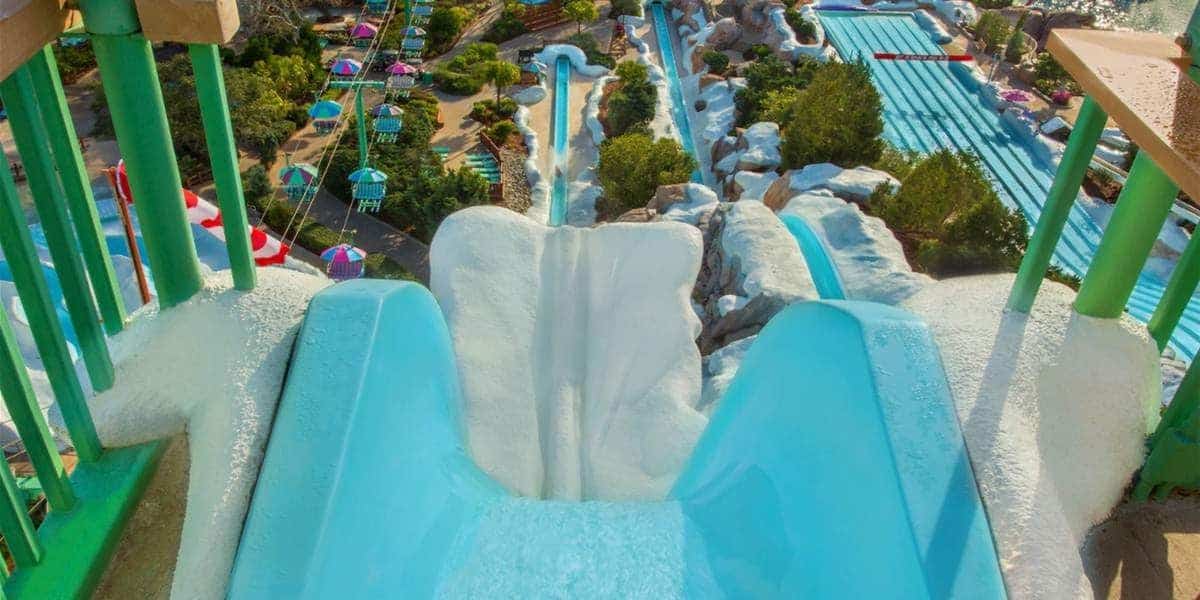 A view from the top of a steep, blue water slide with snow-like decorations on its edges. Green railings frame the slide, which leads down to a vibrant water park at Walt Disney World featuring various slides, pools, and colorful umbrellas. Trees and greenery surround this enchanting Disney attraction.