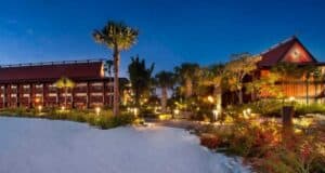 Nighttime view of a tropical resort reminiscent of the Disney Vacation Club Polynesian, with red-roofed buildings and warm lighting. Surrounded by palm trees and lush greenery, a white sandy area graces the foreground. The deep blue sky suggests dusk or early evening.