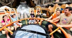 A group of people, including children and adults, are on the Kali River Rapids ride, holding onto the orange safety bars. They are smiling and laughing as they face an upcoming splash of water, set against a rocky backdrop.