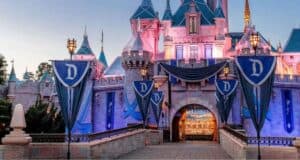 A majestic castle with teal and pink hues at dusk, adorned with blue banners and letter "D" emblems. Warm lights glow invitingly, yet whispers of bad behavior at Disney Parks remind visitors to cherish the enchanting atmosphere. Turrets and spires enhance its fairy-tale appearance as the pathway leads to the entrance.