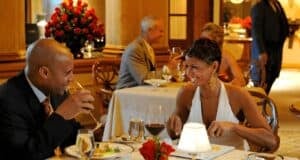 A couple dressed in elegant attire enjoys a romantic meal at one of those enchanting Disney restaurants. The man, wearing a suit, is sipping a drink, while the woman, in a white dress, is smiling and holding a fork. Another couple can be seen dining in the background, with a waiter nearby.