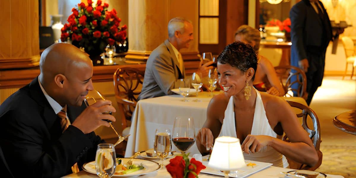 A couple dressed in elegant attire enjoys a romantic meal at one of those enchanting Disney restaurants. The man, wearing a suit, is sipping a drink, while the woman, in a white dress, is smiling and holding a fork. Another couple can be seen dining in the background, with a waiter nearby.