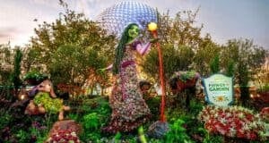 A vibrant floral display features a tall green-skinned figure holding a fruit on a stick beside a stuffed animal character at the Epcot International Flower & Garden Festival, one of the must-dos at Flower & Garden Festival. A geodesic dome structure is visible in the background, with a festival sign in the foreground.