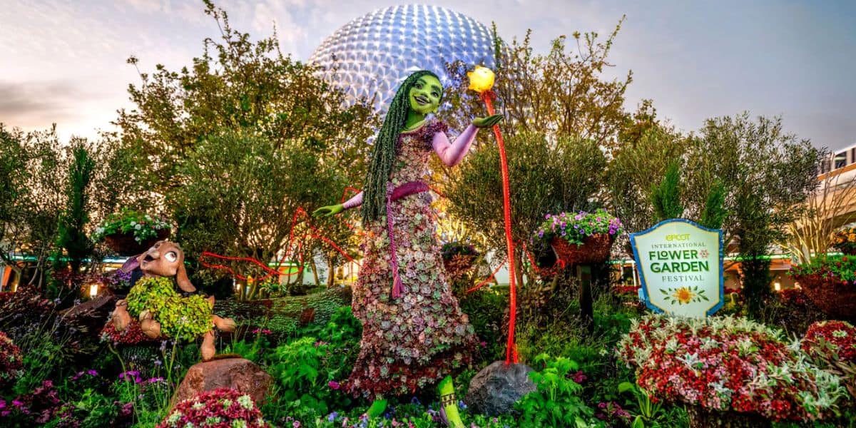 A vibrant floral display features a tall green-skinned figure holding a fruit on a stick beside a stuffed animal character at the Epcot International Flower & Garden Festival, one of the must-dos at Flower & Garden Festival. A geodesic dome structure is visible in the background, with a festival sign in the foreground.