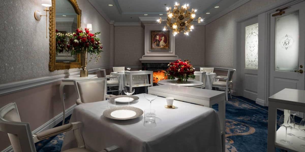 A cozy, elegant dining room features tables with white tablecloths set for two, a beautifully decorated fireplace, ornate chandelier, and floral arrangements. A portrait hangs above the fireplace, and soft lighting creates a warm ambiance—the perfect spot to enjoy My Favorite Eats Money Can Buy at Walt Disney World.