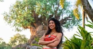 A woman in a colorful costume stands smiling with arms crossed, surrounded by lush greenery reminiscent of Animal Kingdom. A large tree with sprawling branches enhances this Moana-like, vibrant tropical atmosphere.