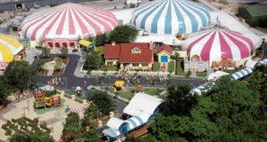Aerial view of a colorful amusement park reminiscent of Mickey's Toontown at Disney World, with pink, blue, and yellow striped circus tents. The park features winding pathways, charming small buildings, and green trees scattered throughout as visitors explore the lively attractions.