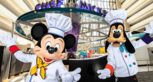Mickey Mouse and Goofy, dressed in chef outfits with white hats and colorful scarves, welcome first-timers to Disney World's vibrant restaurant. The "Chef Mickey's" sign gleams above them in the bright, spacious dining area.
