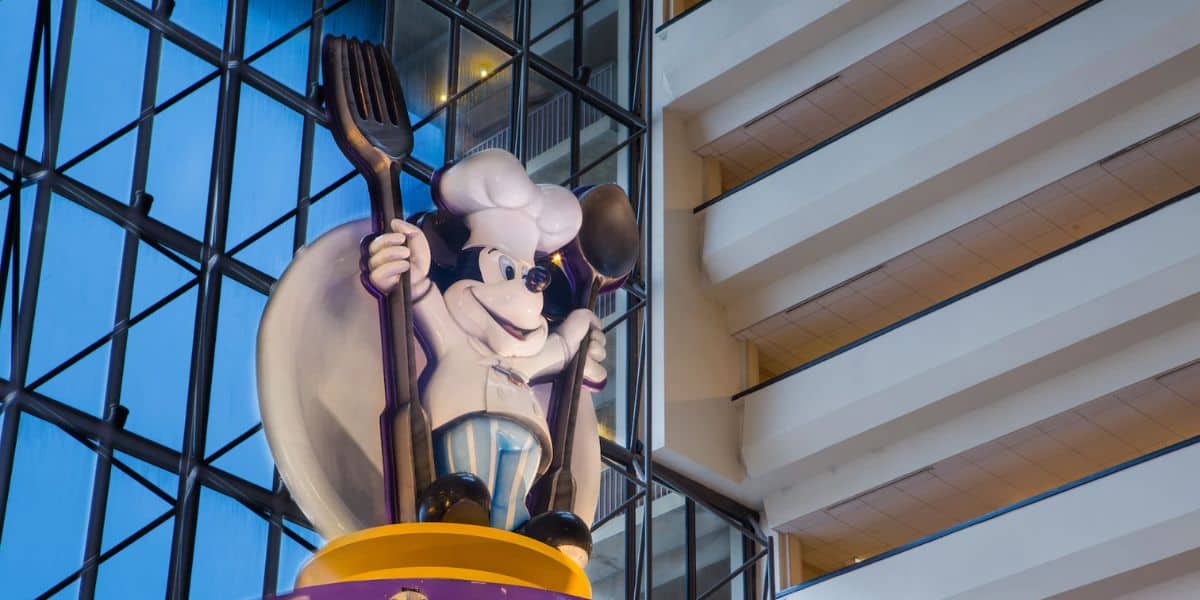 A large statue of a cartoon chef, reminiscent of character experiences at Walt Disney World Resort Hotels, stands inside a building with glass windows. The chef dons a white hat and coat, holding a giant fork and spoon as if to welcome visitors. The interior boasts high ceilings and multiple floors.