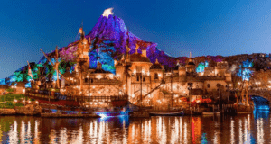 A vibrant night scene of a Disney Parks Worldwide theme park featuring a lit volcano and an ornate, illuminated port. The colorful lights reflect on the water, creating a magical atmosphere with ships and detailed architecture enhancing the enchanting scene.